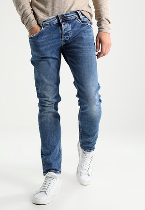 Vaqueros de mezclilla azul con corte recto, que presentan un acabado desgastado y un diseño de cinco bolsillos, combinados con zapatillas blancas.