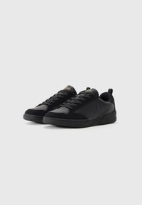 Ein Paar schwarze, niedrige Sneaker mit Schnürdesign und subtiler Logodetail, auf einem schlichten hellgrauen Hintergrund präsentiert.