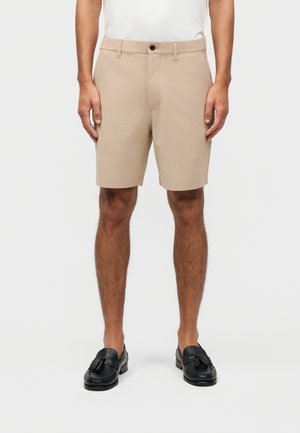 Mężczyzna w beżowych szortach do kolan, białej koszuli i czarnych mokasynach z chwostami, stoi z ramionami swobodnie opuszczonymi wzdłuż ciała.