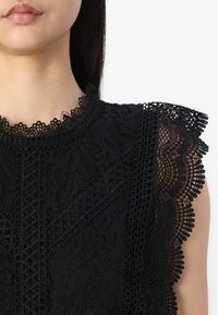 Top en dentelle noire avec des motifs floraux élaborés, un bord en dentelle festonné et des détails texturés autour du col et des manches.