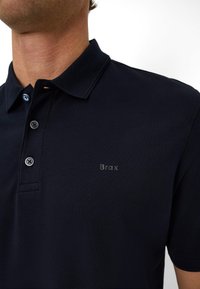 Navy polo shirt made of textured fabric, featuring a buttoned collar, blue buttons, and a subtle "Brax" logo on the chest.