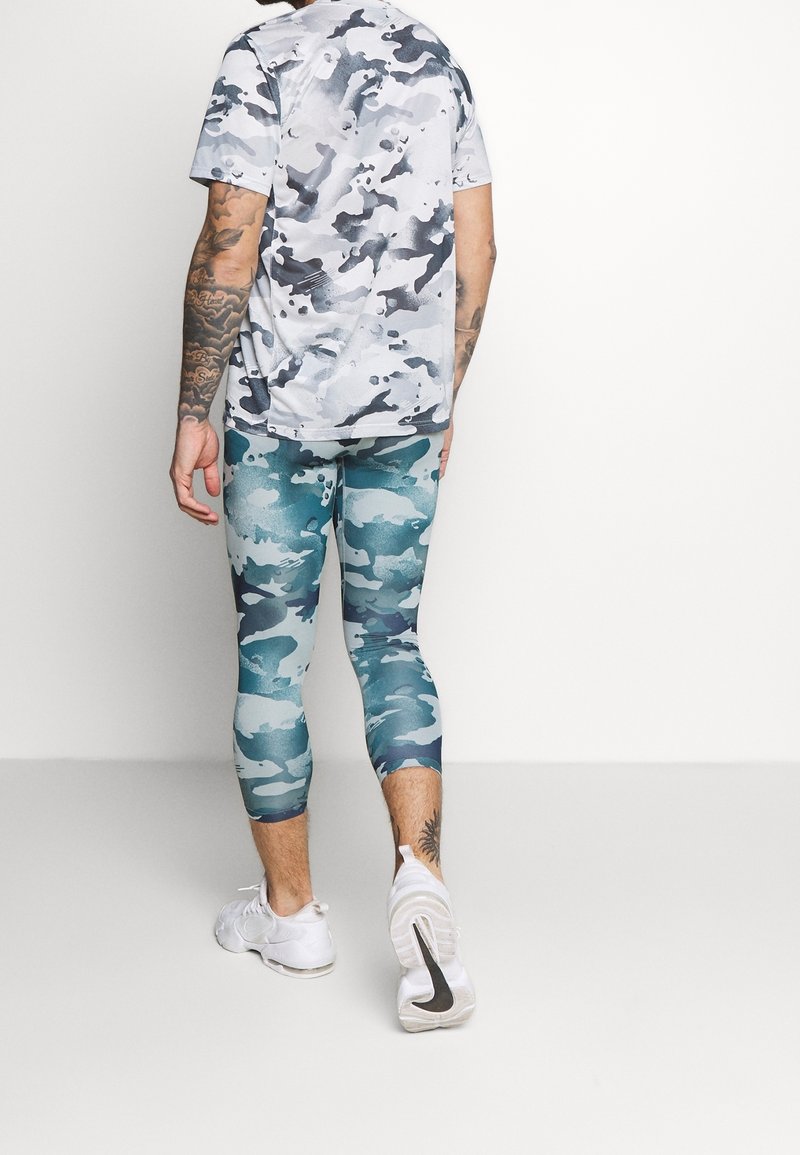 Homme vu de dos portant une chemise camouflage grise et blanche, des leggings courts camouflage bleus, des baskets blanches et des tatouages sur les bras, debout sur un sol gris.