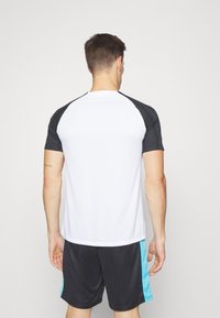 Camiseta atlética blanca con mangas raglán negras, combinada con pantalones cortos negros con acentos turquesa. El tejido parece ligero y suave.