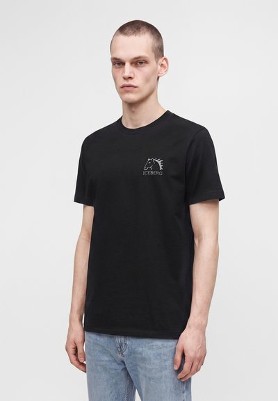 Νέος με κοντά μαλλιά που φοράει μαύρο t-shirt με μικρό λευκό λογότυπο αλόγου και τη λέξη Iceberg στο στήθος, συνδυασμένο με ανοιχτό μπλε τζιν.