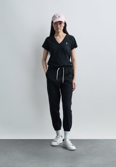 Γυναίκα που στέκεται, φοράει ροζ καπέλο baseball, μαύρο T-shirt με V λαιμόκοψη, μαύρο παντελόνι φόρμας με λευκό κορδόνι, λευκές κάλτσες και λευκά αθλητικά παπούτσια σε γκρι πάτωμα.