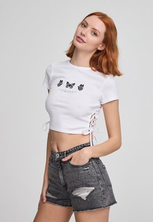 Maglietta bianca cropped con stampe di farfalle nere e dettagli laterali con lacci, abbinata a pantaloni shorts in denim nero strappati a vita alta.