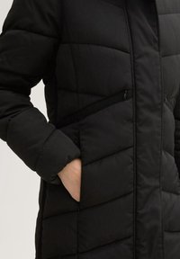 Chaqueta de plumas negra con textura acolchada, bolsillos con cremallera y cuello alto. Presenta un diseño elegante y un ajuste relajado, con una superficie suave.