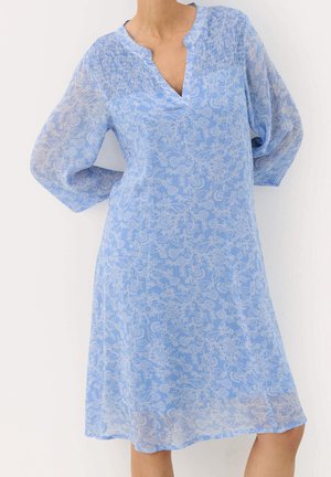 Woman wearing a light blue floral dress with long sleeves and smocked chest detail against a plain white background.