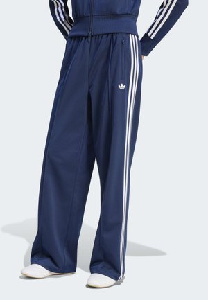 Pantalon de survêtement bleu marine à jambes larges avec bandes blanches sur les côtés et logo, porté avec une veste assortie et des baskets blanches.