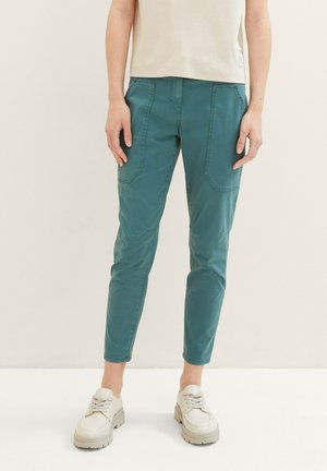 Osoba ubrana w zielonkawo-niebieskie spodnie zwężane z przodu z naszywanymi kieszeniami oraz beżowe buty na sznurowadła, stojąca na jednolitym tle.