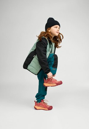 Kind trägt schwarze Mütze, grün-schwarze Jacke, türkisfarbene Hose und bindet auf grauem Hintergrund roten Wanderschuh.