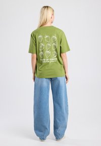 T-shirt en coton vert avec des graphiques de fraises blanches et le texte "Goût de l'Été à Garden Grove" au dos. Coupe lâche, associé à un large jean bleu.