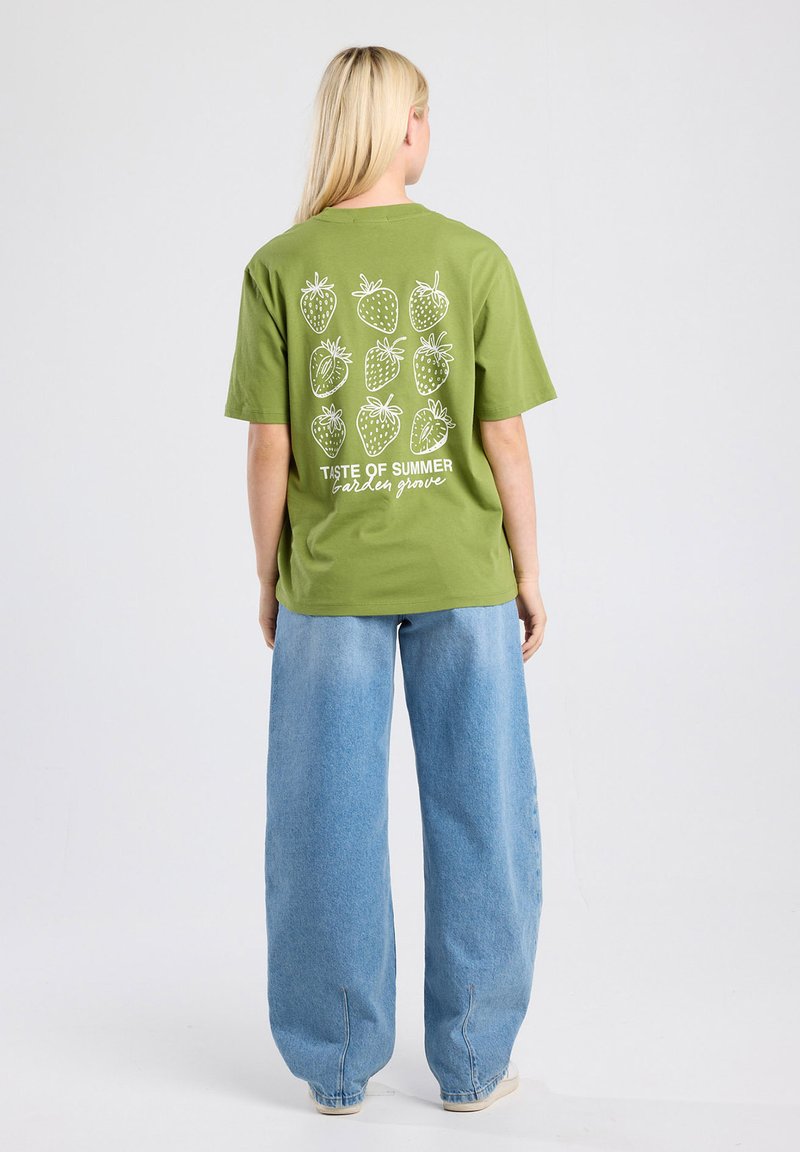 T-shirt en coton vert avec des graphiques de fraises blanches et le texte "Goût de l'Été à Garden Grove" au dos. Coupe lâche, associé à un large jean bleu.