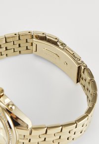 Orologio da polso in metallo dorato con bracciale a maglie, caratterizzato da una finitura lucida, chiusura a scatto e dettagli incisi sul retro.