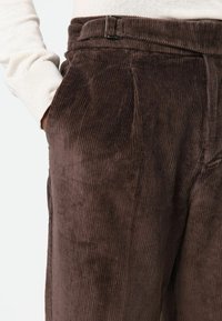 Pantalon en velours côtelé marron avec des nervures verticales, un devant plat et des poches latérales. La ceinture est ornée d'un accent métallique argenté.