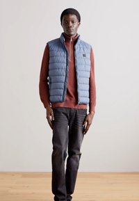 Modrá zateplená vesta přes červenou dlouhou košili, černé džíny, stojící na dřevěné podlaze s jednoduchým bílým pozadím.