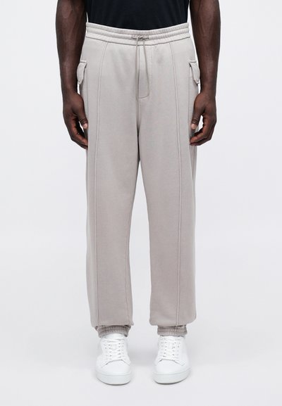 Ανοιχτό γκρι παντελόνι φλις με ελαστική μέση και μανσέτες στους αστραγάλους, που διαθέτει πλαϊνές τσέπες και εμφανείς λεπτομέρειες ραφής. Φοριέται με λευκά αθλητικά παπούτσια.