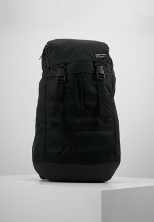 Großer schwarzer Rucksack mit Oberlader, zwei verstellbaren vorderen Schnallenriemen und gepolstertem Boden, ruhend auf einem weißen Ausstellungspodest.