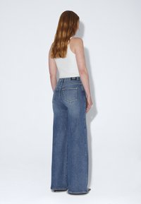 Femme aux longs cheveux bruns portant un débardeur blanc et un jean bleu à jambes larges, vue de dos sur un fond blanc uni.
