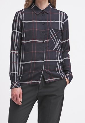 Femme portant une chemise à carreaux noire à manches longues, boutonnée, avec des rayures rouges et blanches et une poche, associée à un pantalon noir.