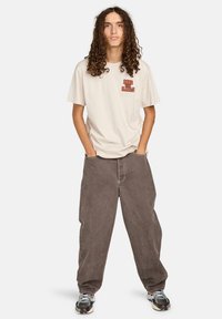 Mladá osoba s dlouhými kudrnatými vlasy stojí s rukama v kapsách, na sobě má béžové tričko "Ride the Rails", volné hnědé kalhoty a tenisky.