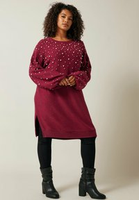 Robe pull oversize bordeaux avec des embellissements en perles, col rond, manches longues, fentes sur les côtés et tissu tricoté texturé. Leggings noirs près du corps et bottes.