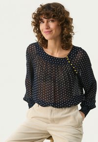 Blouse bleu marine à pois avec un design plissé, des manches longues transparentes et un col rond. Elle est ornée de boutons dorés décoratifs sur l'épaule.