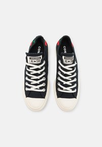 Une paire de baskets Converse All Star noires avec des lacets et des embouts blancs, présentant des motifs de cerises rouges sur la zone du talon.