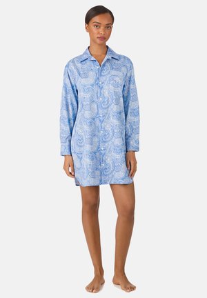 Jonge vrouw die blootsvoets staat en een blauw paisley hemdjurk draagt met lange mouwen, een kraag en een borstzak.