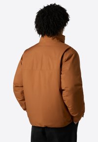 Mann mit lockigem Haar, der eine rostbraune Jacke trägt und mit den Händen in den Taschen steht, dabei von hinten gegen einen einfarbigen hellen Hintergrund gerichtet.