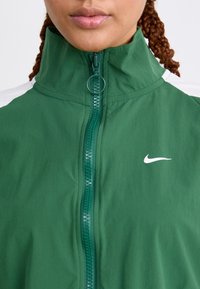 Veste légère verte avec un col haut, fermeture éclair et un logo Nike blanc sur la poitrine. Elle présente une texture lisse et des accents blancs contrastants.