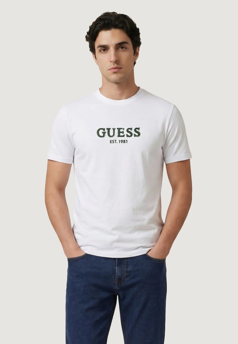 Νεαρός άνδρας που φοράει λευκό T-shirt με κοντό μανίκι και το κείμενο "GUESS EST. 1981" σε σκούρο πράσινο, με τα χέρια στις τσέπες, μπροστά σε μονόχρωμο φόντο.