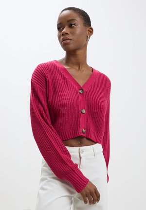 Cardigan court fuchsia avec un col en V, texture côtelée et trois boutons, associé à un pantalon blanc.