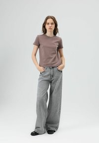 Kortbruin T-shirt met ronde hals, gecombineerd met lichtgrijze jeans met wijde pijpen. De stof lijkt zacht, met zichtbare zakken en stiksels.