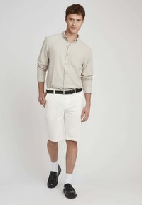 Chemise beige clair à boutons avec une poche, associée à un short blanc et des chaussures noires. Le mannequin se tient avec assurance, mettant en valeur l'ajustement de la tenue.