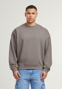 Sweatshirt gris à col rond avec manches longues, fabriqué en tissu doux. Coupe légèrement oversize avec des poignets et un ourlet côtelés. Porté avec un jean bleu clair.