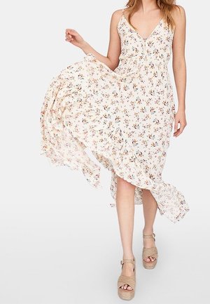 Vrouw draagt een vloeiende witte mouwloze jurk met bloemenprint en beige platformzandalen, houdt de jurk vast terwijl deze wapperend beweegt.