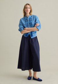 Blusa azzurro chiaro con ricami blu scuro, abbinata a una gonna midi blu navy e ballerine blu. La blusa ha un collo rotondo e bottoni.