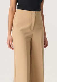 Pantaloni larghi beige in un tessuto morbido e liscio con una vestibilità sartoriale, caratterizzati da un davanti piatto e cuciture laterali precise per una silhouette pulita.