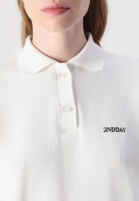 Biela polo košeľa vyrobená z textúrovanej látky s klasickým golierom, s tromi gombíkmi a malým čiernym logom "2NDDAY" na hrudi.