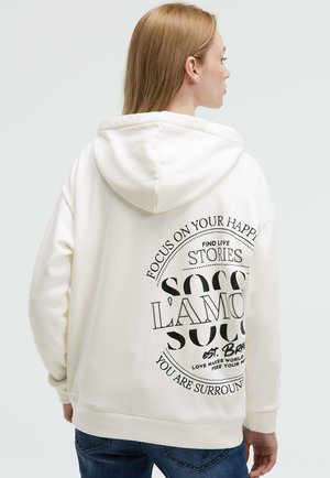 Woman with light brown hair wearing a white hoodie with black circular text and blue jeans, facing away against a plain background.