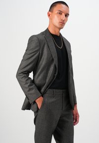Sacou gri în țesătură herringbone, cu o croială slim, asociat cu o cămașă neagră. Are un închidere cu un singur buton și o textură subtilă.