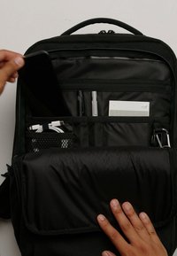 Sac à dos noir avec plusieurs compartiments ; comprend une poche zippée, une section en maille et des emplacements pour stylos et un appareil mobile. Texture lisse.