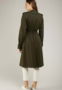 Manteau trench vert olive avec une taille ceinturée, un grand col et des sangles de manche ajustables. Texture lisse et design longueur genoux.