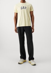 Světlé žluté bavlněné tričko s nápisem "GAP" v tmavě modrých písmech, doplněné černými kalhotami a šedými teniskami se žlutými detaily.