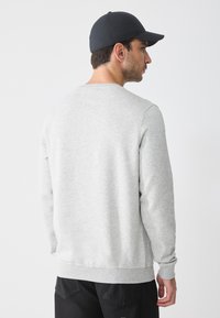 Sudadera gris de manga larga y cuello redondo, hecha de una tela suave, combinada con una gorra negra, vista desde la parte de atrás.
