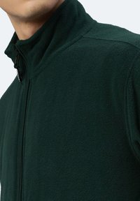Dunkelgrüne Fleecejacke mit hohem Kragen, durchgehendem Reißverschluss und strukturiertem Stoff. Verfügt über ein kleines Markenetikett in der Nähe des Reißverschlusses.