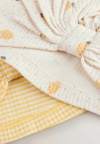 Sombrero de bebé suave en crema y amarillo con detalle de lazo, que presenta pequeños patrones de puntos y círculos y tela interior a rayas amarillas.