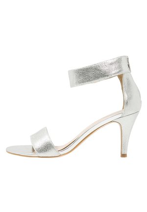 Sandał na wysokim obcasie w srebrnym metalicznym kolorze, z paskiem wokół kostki, odkrytymi palcami i teksturowaną powierzchnią. Szpilka o smukłym kształcie z minimalistycznymi dodatkami.