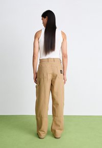 Beige cargo pants made of thick cotton, featuring large side pockets, a back label, and a relaxed fit, worn with a white tank top.